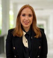 Kathryn Williams, eine Frau mit glattem rotem Haar, die einen schwarzen Blazer über einem schwarz-weißen Hemd trägt, steht in einem hell erleuchteten Korridor und lächelt in die Kamera.
