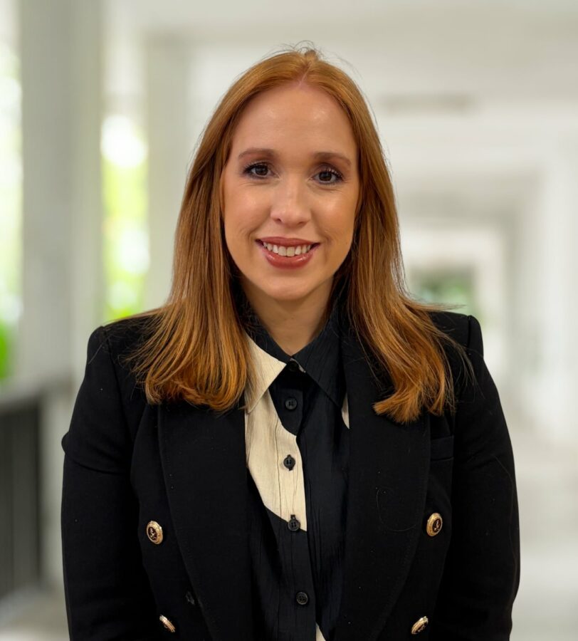Kathryn Williams, eine Frau mit glattem rotem Haar, die einen schwarzen Blazer über einem schwarz-weißen Hemd trägt, steht in einem hell erleuchteten Korridor und lächelt in die Kamera.