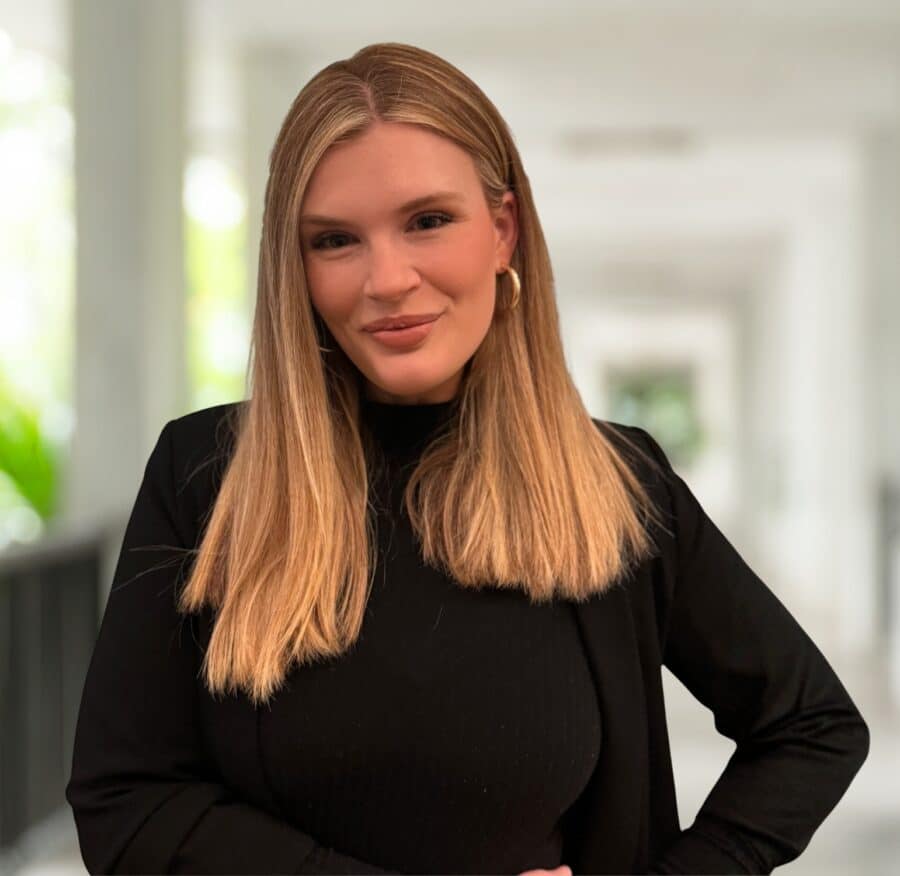 Claire Butcher, mit glattem blondem Haar, steht in einem schwarzen Oberteil und einem Blazer im Haus und schaut mit einem leichten Lächeln in die Kamera.