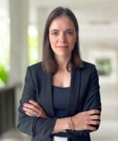 Claudia Lemke, eine Frau mit glattem braunem Haar, die einen schwarzen Blazer und ein schwarzes Oberteil trägt, steht in einem Raum, die Arme verschränkt, und blickt direkt in die Kamera. Der Hintergrund ist leicht unscharf.
