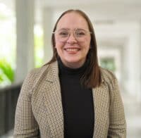 Haley Gibson, eine Person mit Brille und glattem braunem Haar, die einen karierten Blazer über einem schwarzen Rollkragenpullover trägt, lächelt, während sie in einem hellen Korridor steht.