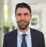 Manuel Preg, gekleidet in einen dunklen Anzug, ein hellblaues Hemd und eine gemusterte Krawatte, lächelt, während er in einem hellen, modernen Innenraum vor einem unscharfen Hintergrund steht.