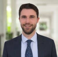 Manuel Preg, ein Mann mit kurzen braunen Haaren und einem Bart, der einen dunklen Anzug, ein hellblaues Hemd und eine gemusterte Krawatte trägt, steht in einem gut beleuchteten Korridor und lächelt in die Kamera.