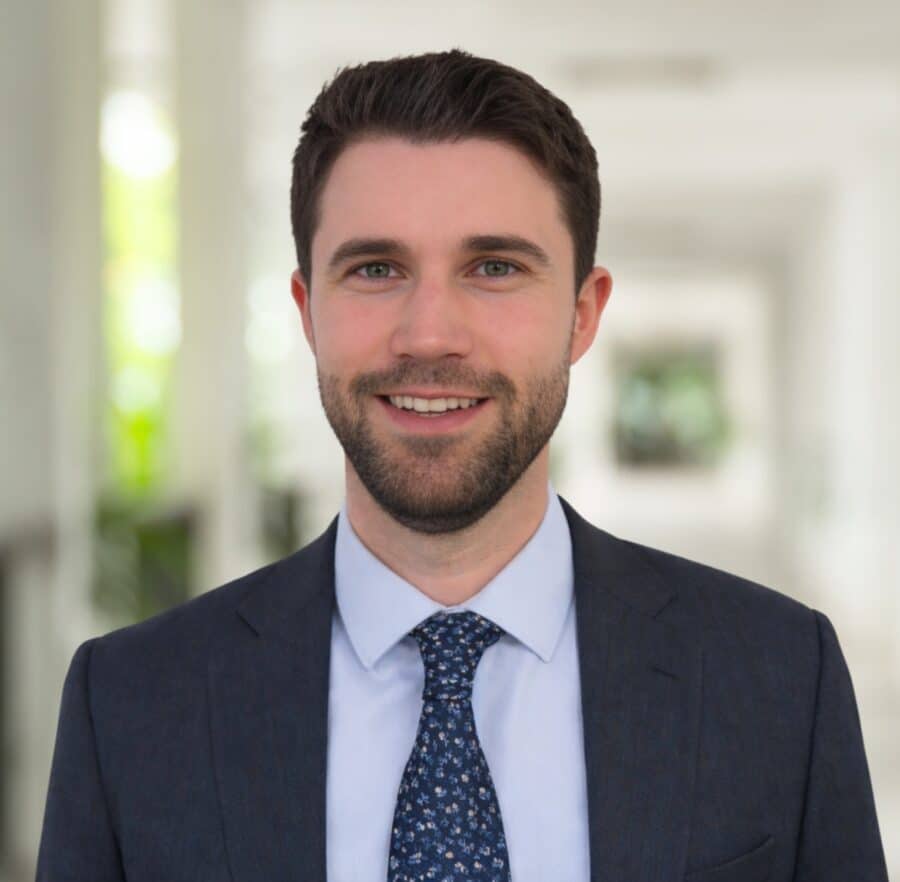 Manuel Preg, ein Mann mit kurzen braunen Haaren und einem Bart, der einen dunklen Anzug, ein hellblaues Hemd und eine gemusterte Krawatte trägt, steht in einem gut beleuchteten Korridor und lächelt in die Kamera.