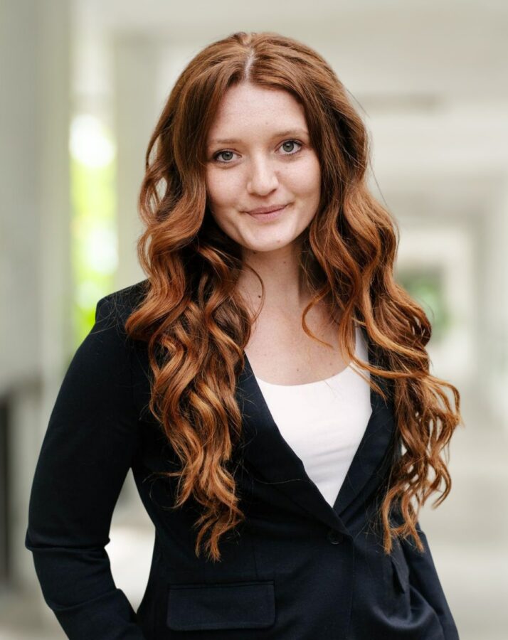 Eine Frau mit langen, gewellten roten Haaren, die einen schwarzen Blazer und ein weißes Oberteil trägt, steht in einem hellen Flur und blickt in die Kamera - ein Bild, das an Adam Nuzies typischen Stil erinnert.