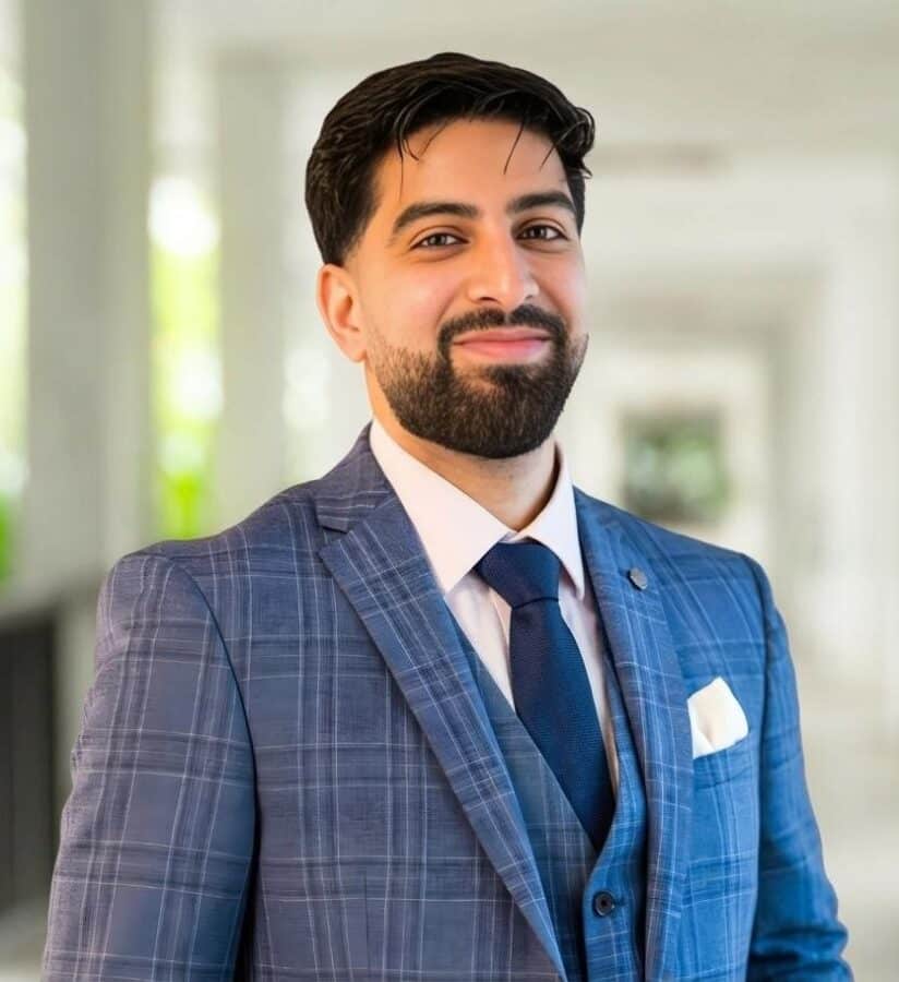 Ein Mann mit dunklem Haar und Bart trägt einen blau karierten Anzug, ein weißes Hemd und eine marineblaue Krawatte. Er steht in einem hellen, verschwommenen Innenkorridor, der an Sahar Akhtars anspruchsvollen Stil erinnert.