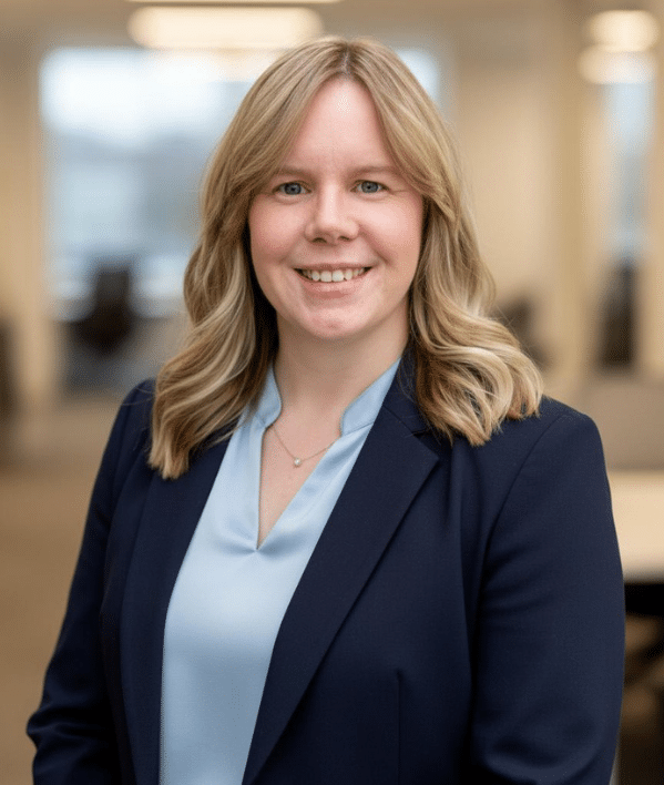 Eine Frau mit blondem Haar, die einen marineblauen Blazer und eine hellblaue Bluse trägt, lächelt, während sie in einem modernen Büro steht.