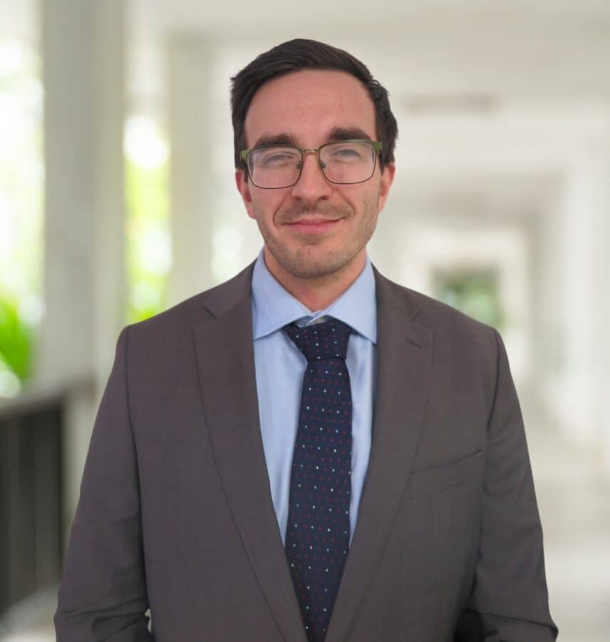 Ein Mann mit Brille und im Anzug steht drinnen in einem Flur, blickt mit ausdruckslosem Gesicht in die Kamera.
