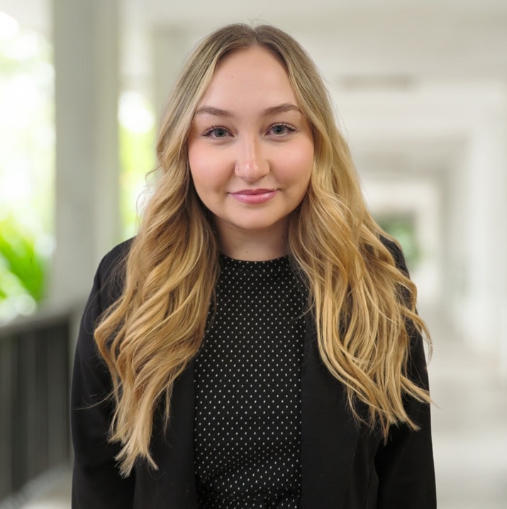 Eine Frau mit langen blonden Haaren, die einen schwarzen Blazer und ein gepunktetes Oberteil trägt, steht in einem hellen, verschwommenen Flur.