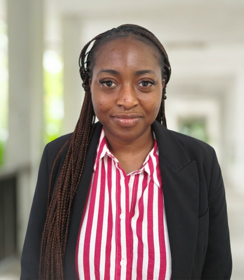 Feyisayo Adetayo, mit langen Zöpfen, trägt einen schwarzen Blazer über einem rot-weiß gestreiften Hemd und steht mit neutraler Miene in einem hellen Korridor.