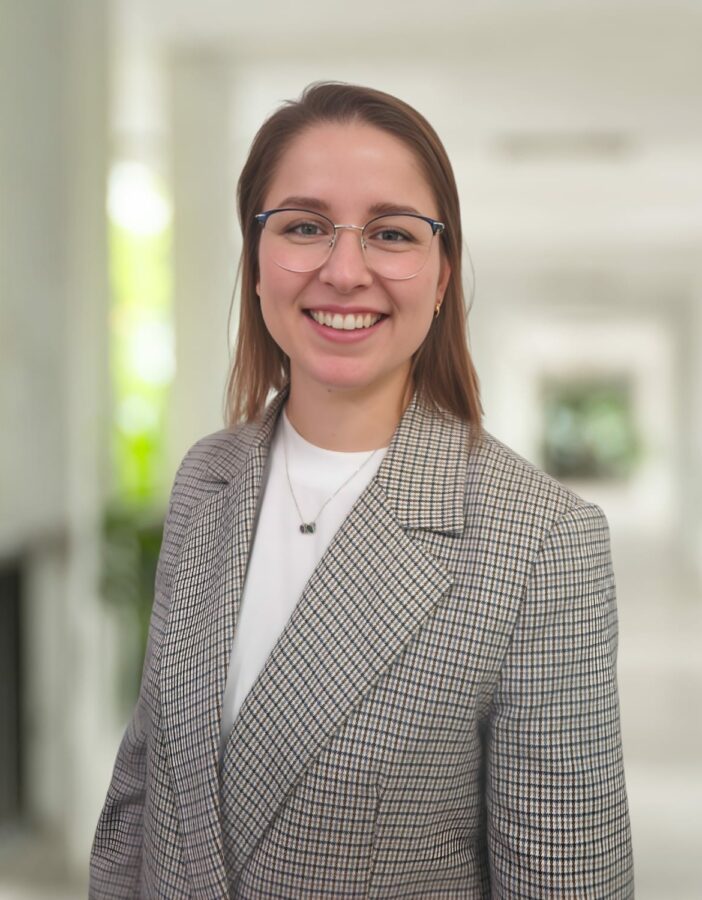 Eine Frau, die eine Brille und einen karierten Blazer trägt, lächelt, während sie in einem hellen Hausflur mit unscharfem Hintergrund steht.