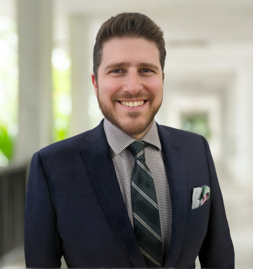 Ein Mann in einem dunklen Anzug, einem karierten Hemd und einer gestreiften Krawatte steht lächelnd auf einem hellen, überdachten Gehweg mit verschwommenem Grün im Hintergrund.