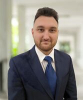 Ein Mann im blauen Anzug und mit Krawatte steht auf einem hellen, überdachten Gehweg mit Grünpflanzen im Hintergrund.