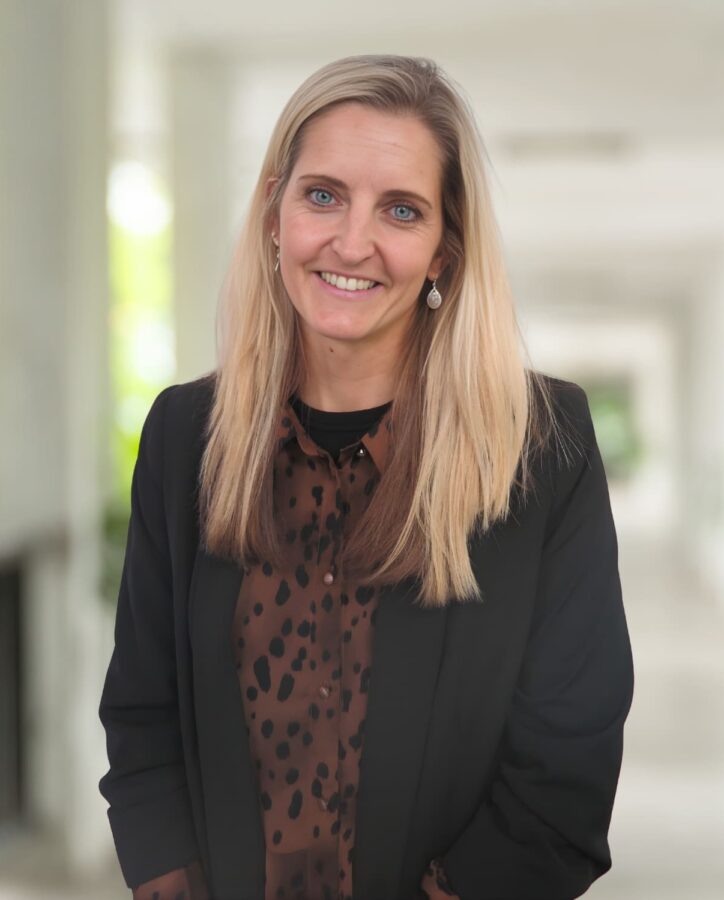 Eine Frau mit langen blonden Haaren, die einen schwarzen Blazer und eine braun gemusterte Bluse trägt, steht in einem sonnenbeschienenen Korridor und lächelt in die Kamera.