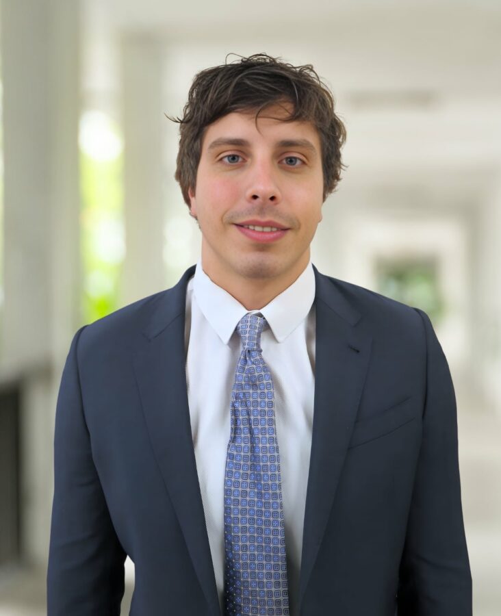 Pat Cunningham, im dunklen Anzug und mit gemusterter Krawatte, steht in einem hellen Korridor im Freien mit Säulen und Grünpflanzen im Hintergrund.