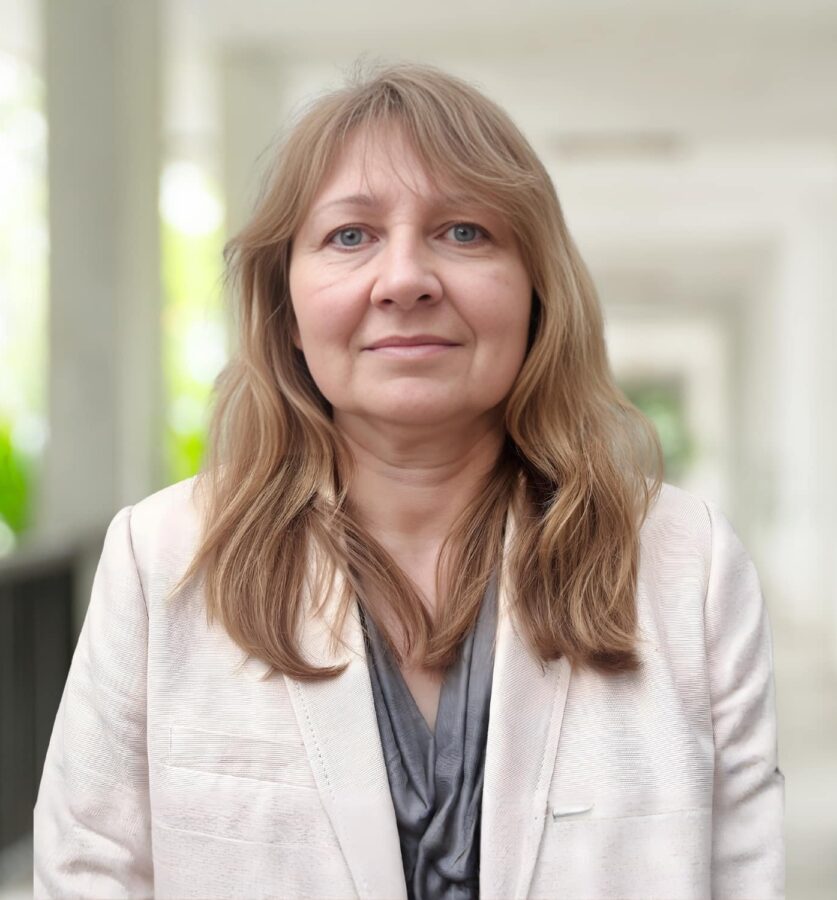Eine Frau mit hellbraunem Haar, die einen hellen Blazer trägt, steht in einem hellen, überdachten Gehweg im Freien mit Grün im Hintergrund.