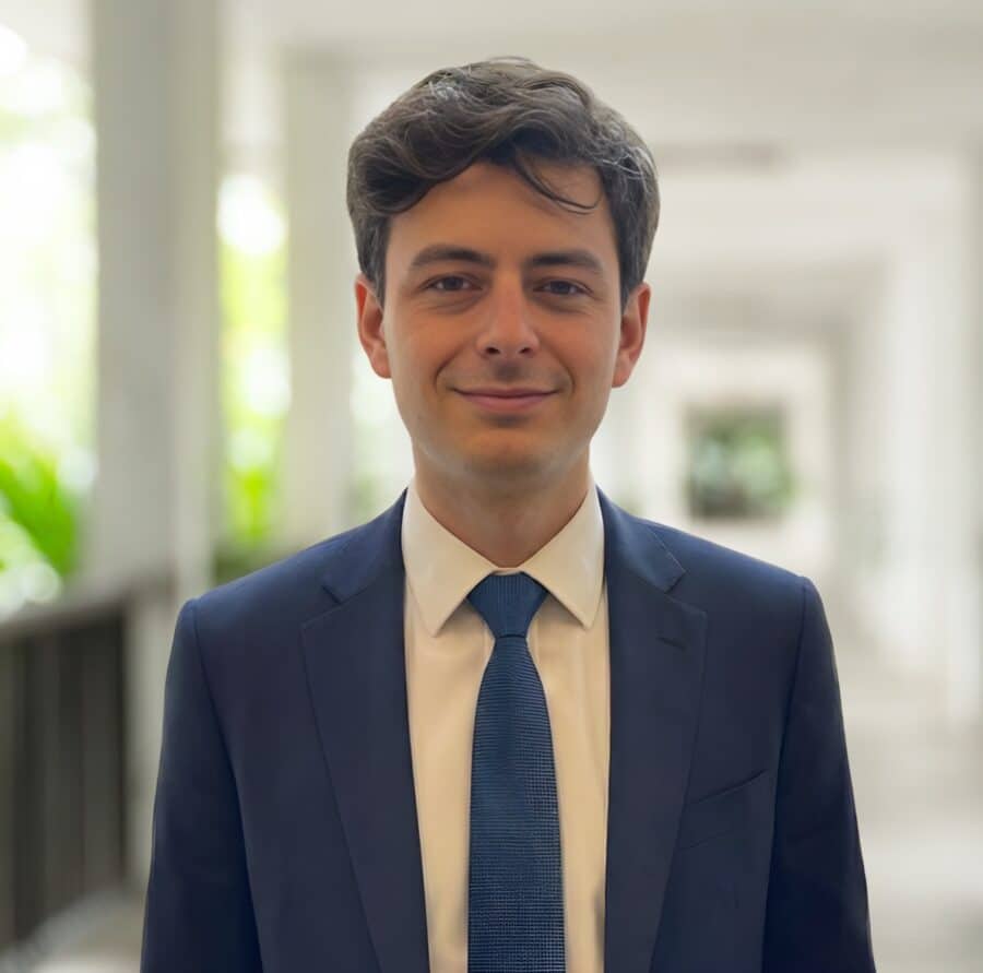 Ein Mann in blauem Anzug und Krawatte steht in einem hellen Korridor mit verschwommenen Säulen und Grünpflanzen im Hintergrund.