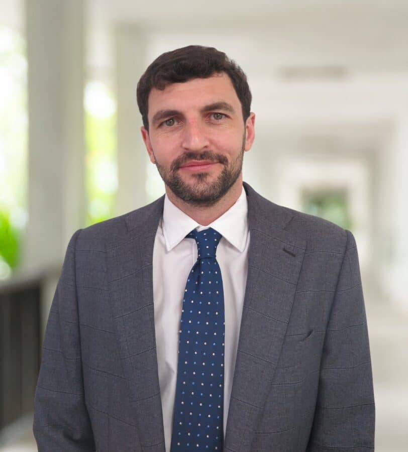 Ein Mann im grauen Anzug und mit blauer Krawatte steht in einem hellen Flur und schaut mit neutralem Blick in die Kamera.