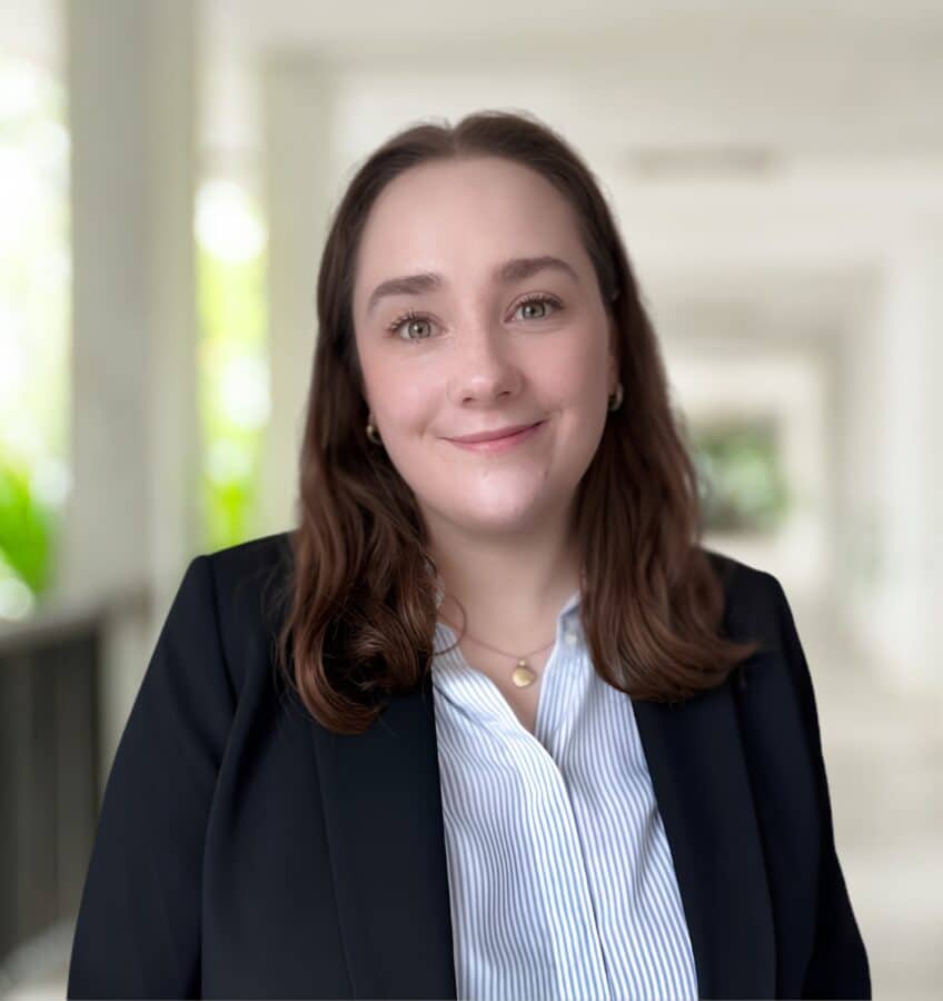 Sophia Minns, eine Frau mit braunen Haaren, die einen dunklen Blazer und ein gestreiftes Hemd trägt, steht in einem hellen, verschwommenen Flur und lächelt in die Kamera.