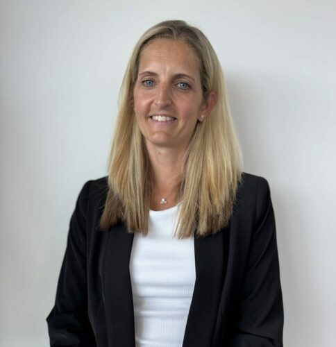 A woman with long blonde hair, wearing a black blazer over a white top, stands in front of a plain white background, smiling at the camera.