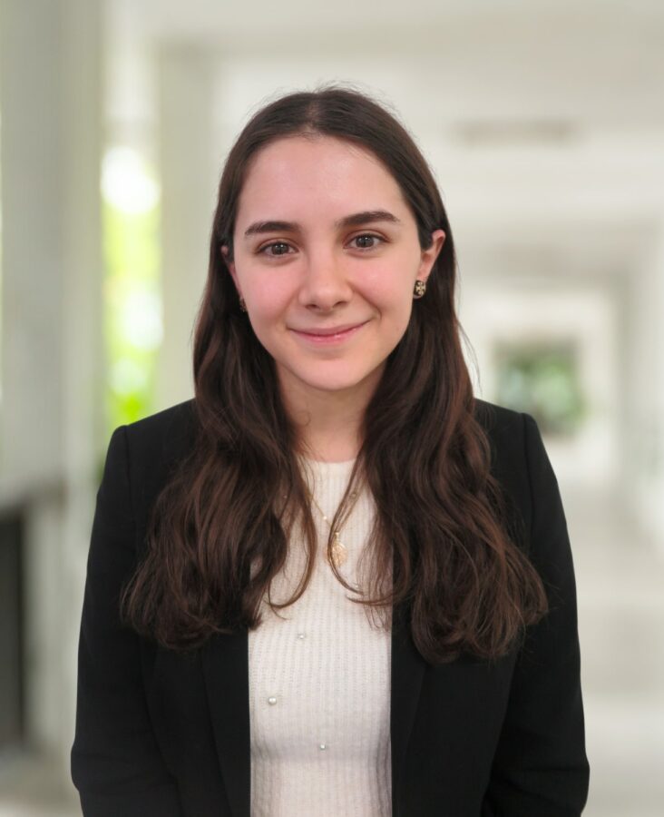 Eine junge Frau mit langen braunen Haaren, die einen schwarzen Blazer und ein weißes Oberteil trägt, steht in einem hellen, verschwommenen Flur und lächelt in die Kamera.