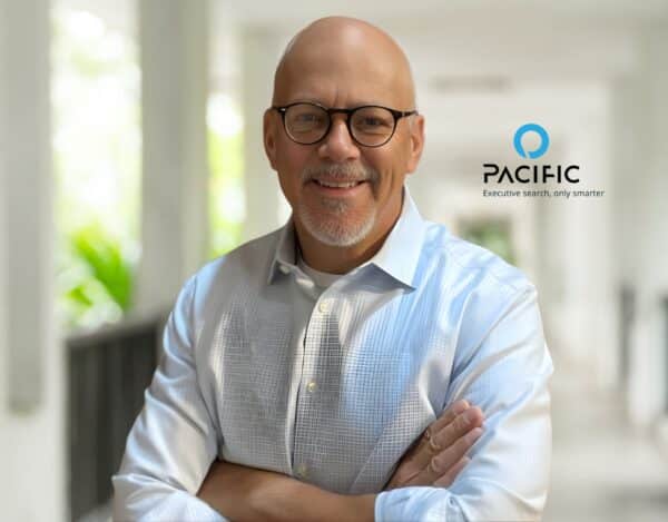 A man wearing glasses and a light blue shirt stands with arms crossed in a bright corridor. The Pacific company logo, slogan, and Advisory Board mention appear beside him.