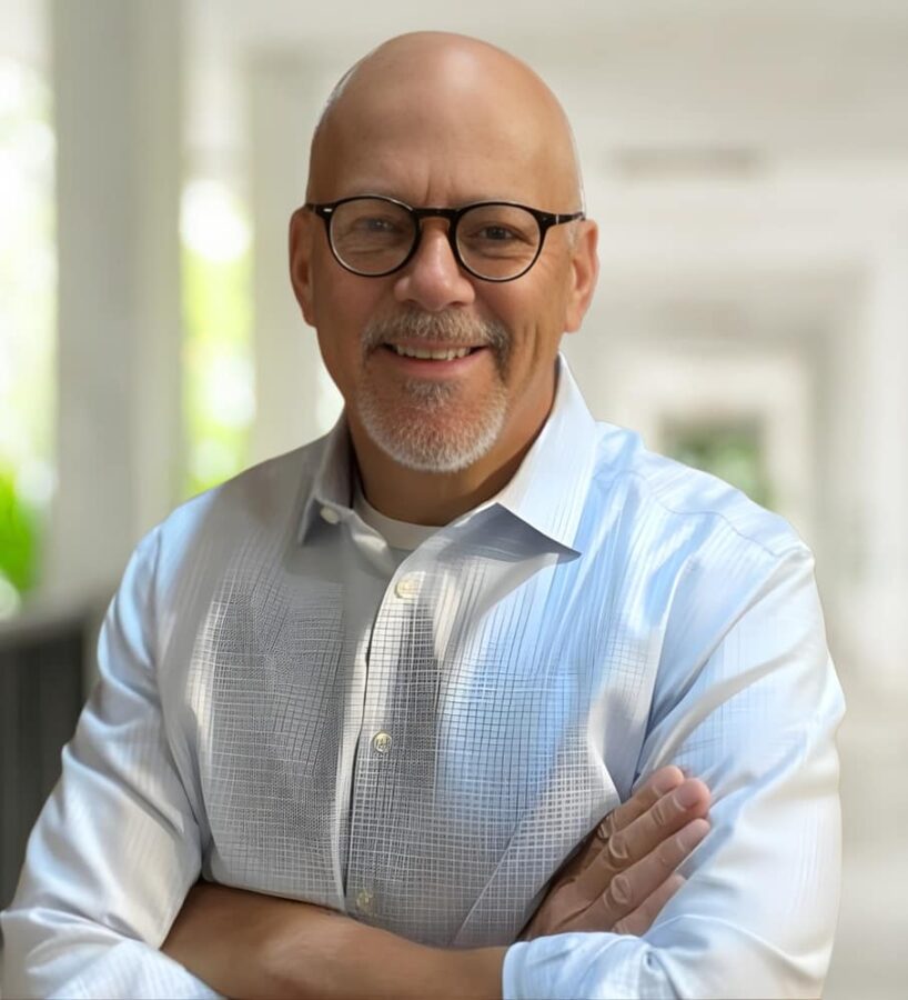 A man with glasses and a short beard smiles with arms crossed, wearing a light blue shirt, standing in a brightly lit corridor.