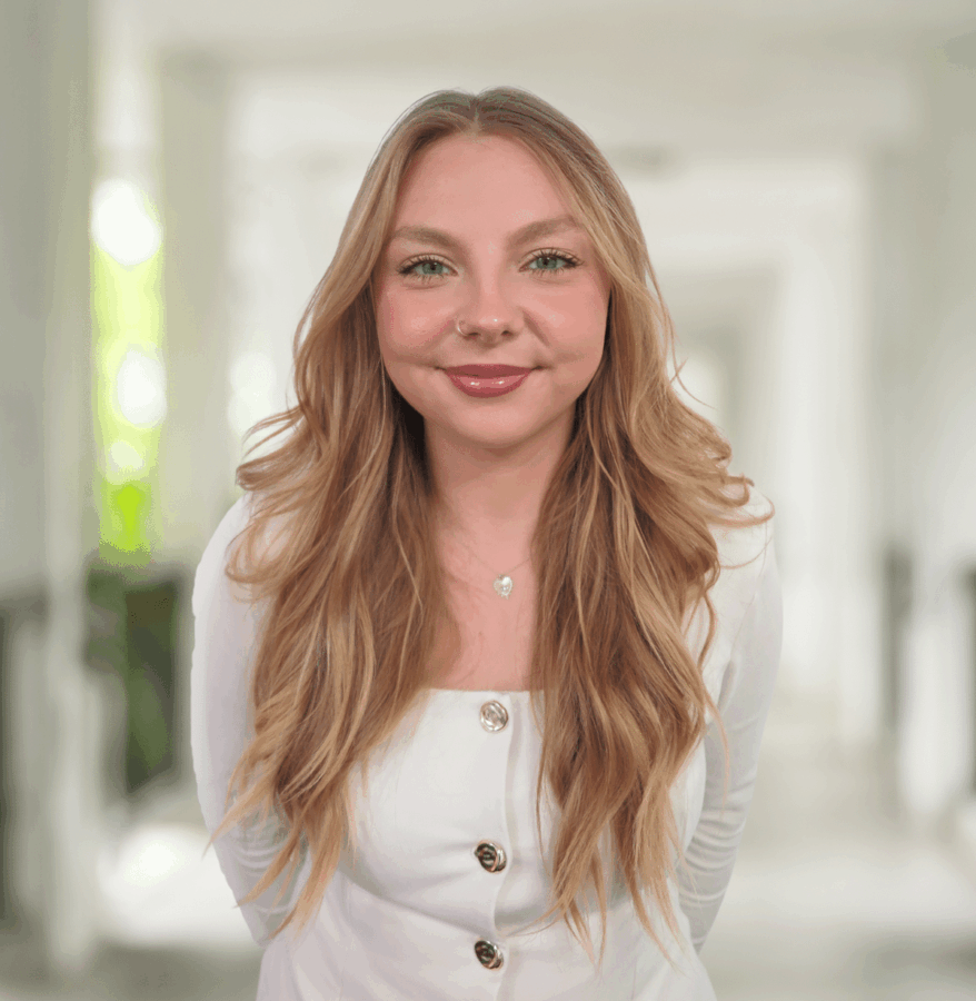 A young woman with long blonde hair, wearing a white top with buttons, stands indoors in a bright corridor, smiling at the camera.