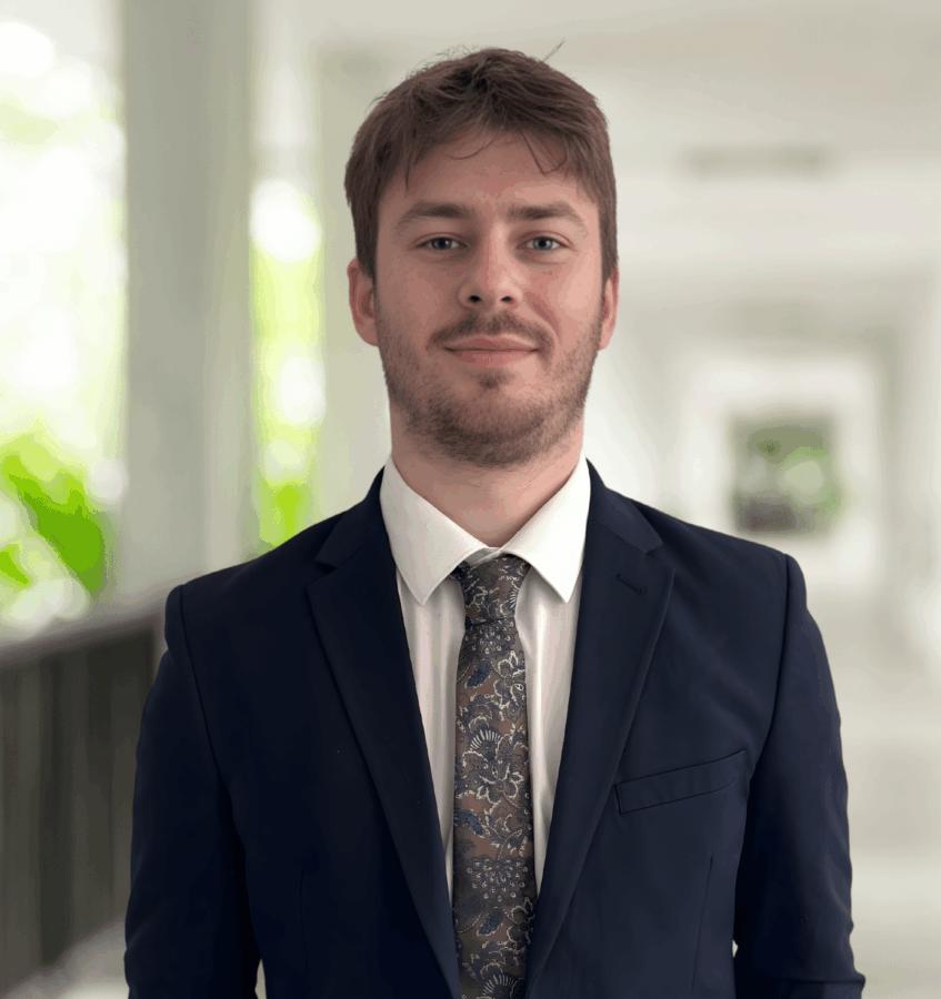 A young man in a dark suit, white shirt, and patterned tie stands in a bright corridor with blurred background and greenery visible.
