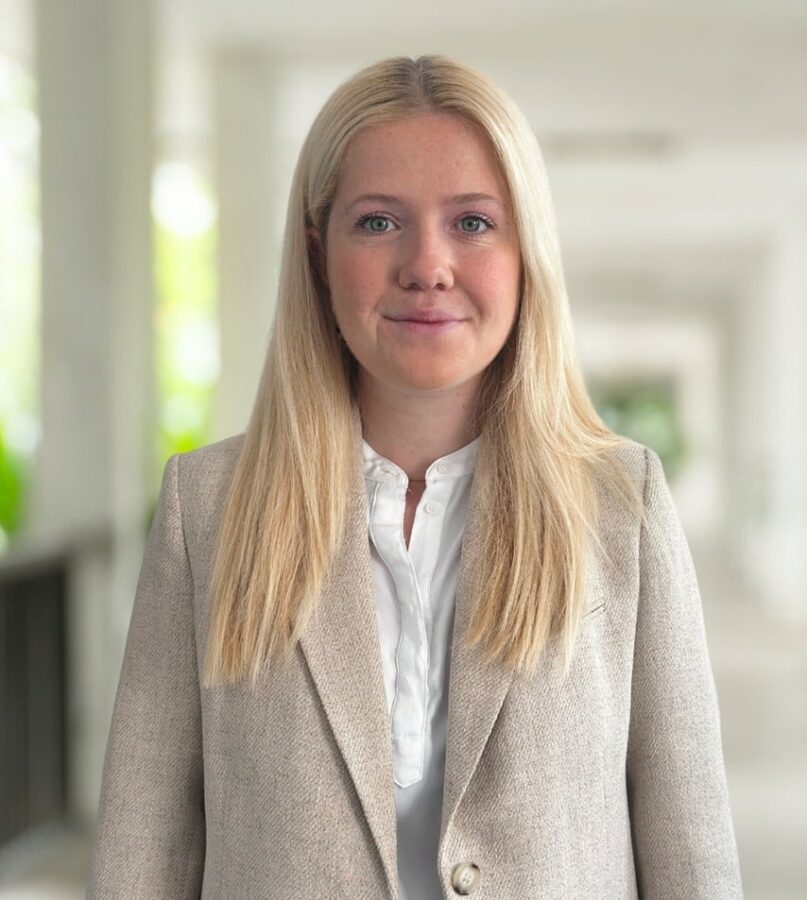 A woman with long blonde hair, wearing a beige blazer over a white blouse, stands in a bright, blurred indoor corridor.