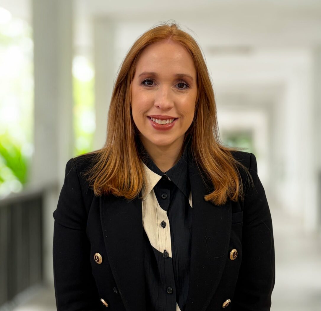 Eine Frau mit schulterlangen roten Haaren, die eine schwarze Jacke und eine cremefarbene Bluse trägt, steht in einem hellen Flur mit verschwommenen Säulen - eine inspirierende Umgebung, um sich unserem dynamischen Team anzuschließen.