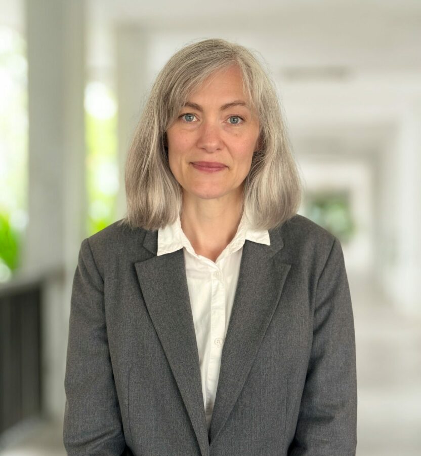 A woman with grey hair in a grey blazer and white shirt stands in a bright corridor, looking at the camera with a neutral expression.