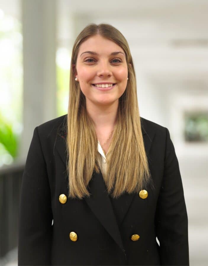 Eine Frau mit langen blonden Haaren, die einen schwarzen Blazer mit goldenen Knöpfen trägt, steht in einem Raum und lächelt in die Kamera.