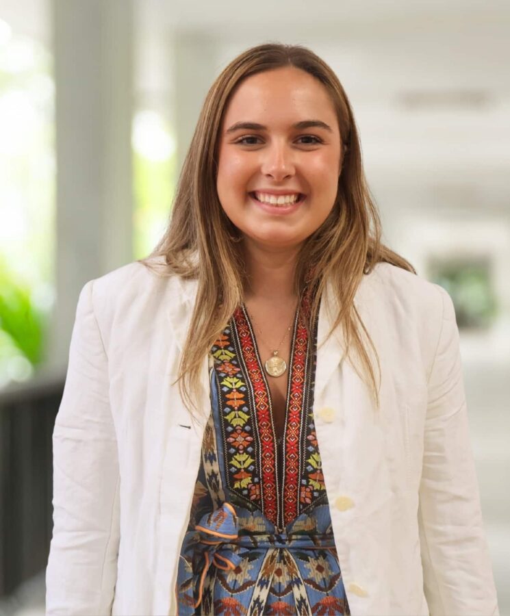 A woman stands indoors, smiling at the camera. She wears a white jacket over a patterned dress and a necklace. The background is softly blurred.